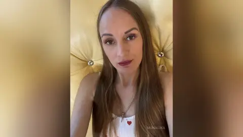 Young woman with long brown hair wearing a white tank top and gold necklace in a cozy indoor setting.