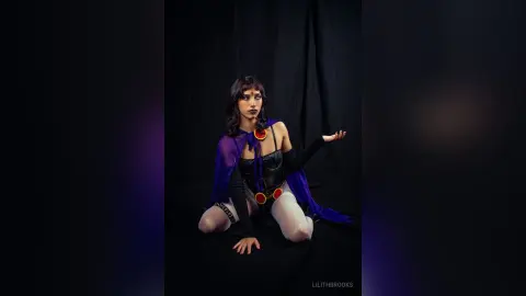 LilithBrooks poses in a black and purple outfit with sheer sleeves and red accents against a dark background.