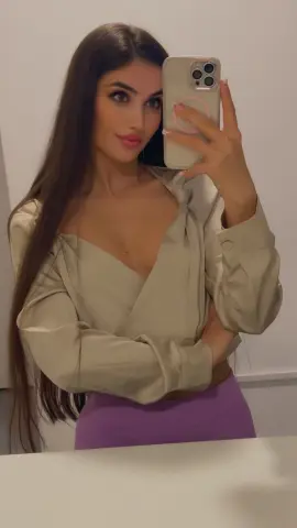 Young woman with long dark hair wearing a beige wrap blouse and purple skirt in a bright indoor setting.