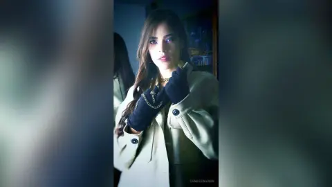 Moody portrait of a woman in a beige trench coat and black gloves posing in dim indoor lighting.