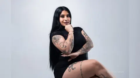 BellaMonrow poses in a black dress, showing off her long black hair and detailed arm and leg tattoos against a plain white background.