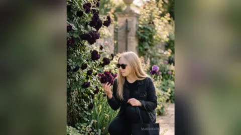 Blonde woman in a black jacket and sunglasses holding purple flowers in a lush, dreamy summer garden.