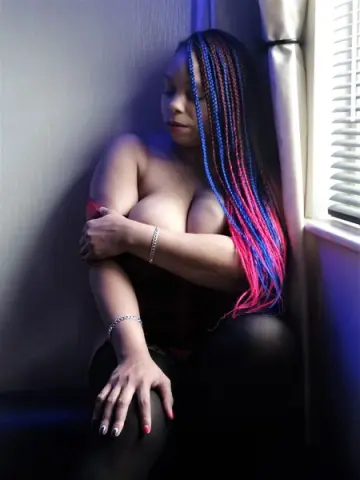 Moody portrait of a woman with colorful blue and pink braids wearing black clothing by a window.