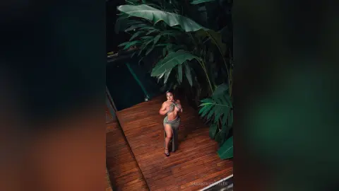 Fashionable woman in a sage green cutout maxi dress standing on a wooden deck surrounded by lush tropical plants.