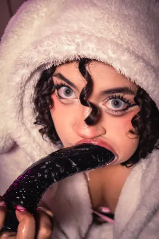 Young woman with curly hair in a cozy white fuzzy hoodie eating a dark purple eggplant in a soft mood.