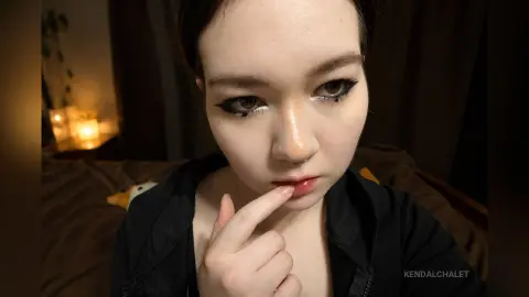 Young woman with dramatic winged eyeliner and black outfit in a cozy, dimly lit room.
