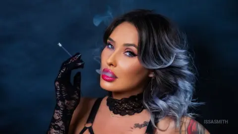 Glamorous woman with blue hair and lace gloves smoking a cigarette against a moody dark blue background.