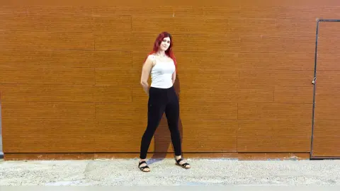 Red-haired woman in a white tank top and black leggings standing against a rustic wooden wall.