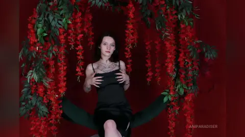 Moody portrait of a woman in a black slip dress sitting amidst hanging red flowers against a red backdrop.