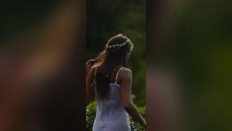 Dreamy portrait of a woman in a white summer dress wearing a flower crown in a lush green garden.