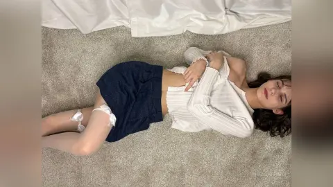 Young woman in a white long-sleeve top and navy skirt lying on a carpeted floor in a relaxed mood.