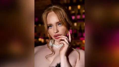 Elegant woman in a black lace outfit and pearl necklace posing in a moody, dimly lit lounge setting.