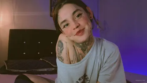 Smiling young woman with neck tattoos wearing a casual grey t-shirt in a dimly lit bedroom setting.