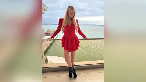 Blonde woman in a stylish red mini dress posing on a balcony overlooking the ocean at sunset.