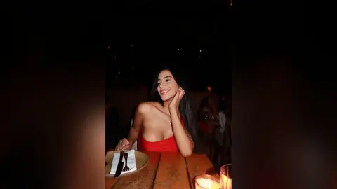 Alison smiles while sitting at a wooden table in a red top, resting her hand near her long dark hair.