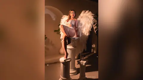 HillaryLewis poses in white lace lingerie and large white angel wings, sitting on a white pillar in a dimly lit room.