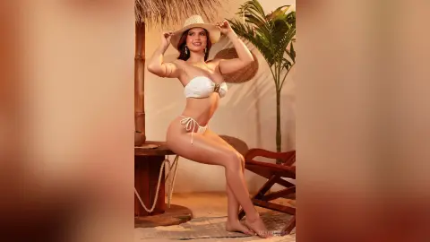 Smiling woman in white bikini and sun hat posing in a tropical resort setting.