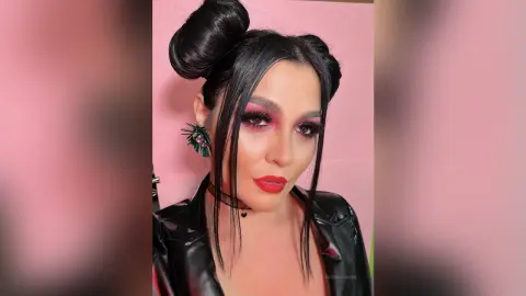 Woman with dramatic smoky eye makeup and black leather jacket posing against a pink background.