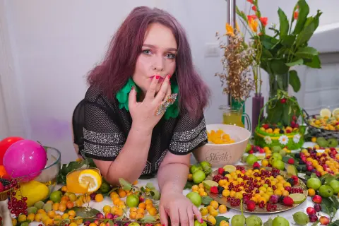 Young woman with purple hair in a black mesh top posing by a table of colorful tropical fruits.