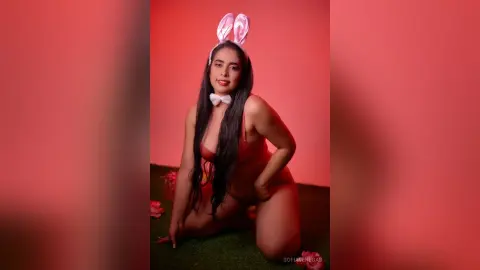Woman in red bunny outfit and white bow tie sitting on grass against a pink background.