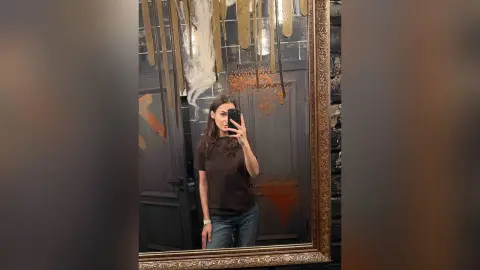 Young woman in a brown t-shirt and jeans taking a mirror selfie in a moody, dimly lit room.