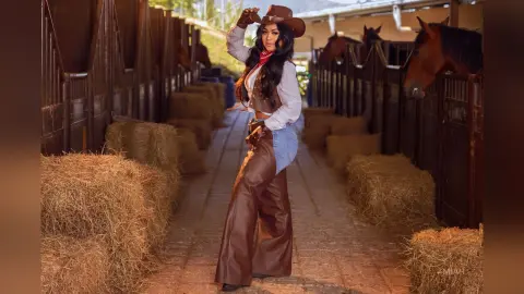 Stylish cowgirl in brown leather chaps and cowboy hat posing in a rustic horse stable with hay bales.