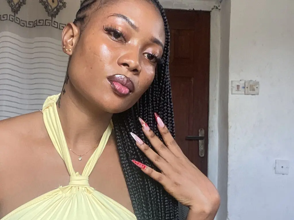 Beautiful Black woman with long braids and long nails wearing a yellow halter top in a cozy indoor setting.