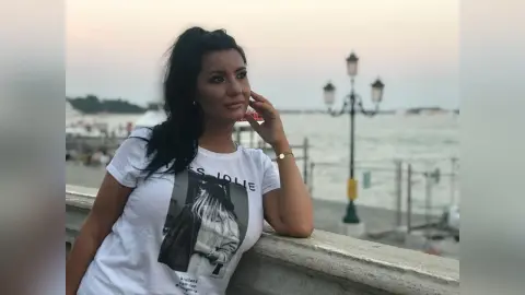 MissyJolie leans against a stone ledge overlooking the ocean, wearing a white graphic t-shirt with her name on it.