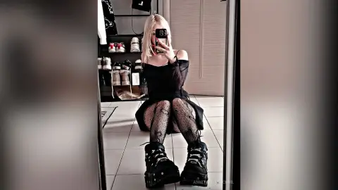 Blonde woman in black dress and fishnet tights posing in a mirror for a moody aesthetic selfie.