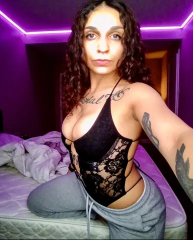 Woman with curly hair in a black lace bodysuit sitting on a bed under purple mood lighting.