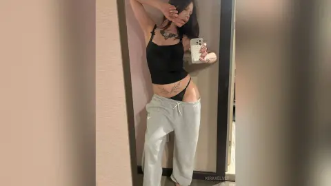 Young woman in a black tank top and grey sweatpants taking a mirror selfie in a modern room.