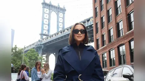 Stylish woman in a navy blue trench coat and sunglasses standing before the Manhattan Bridge in New York City.