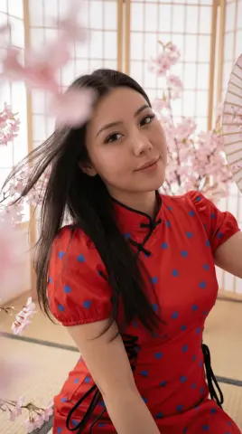 Smiling young woman in a red floral cheongsam dress posing against pink cherry blossoms in a serene setting.