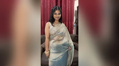 Sejuti poses in a light blue silk saree and silver necklace, standing in front of red curtains with her long dark hair down.