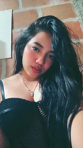 Young woman with long dark hair wearing a black mesh top and silver necklace against a brick wall.