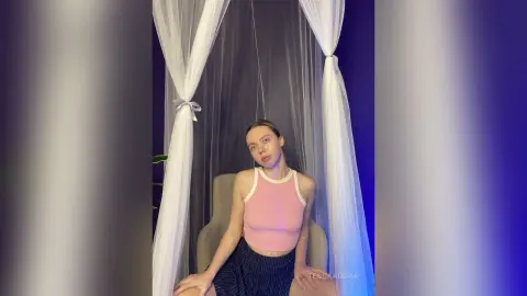 Young woman in a pink tank top sitting under white sheer curtains in a moody, dimly lit room.
