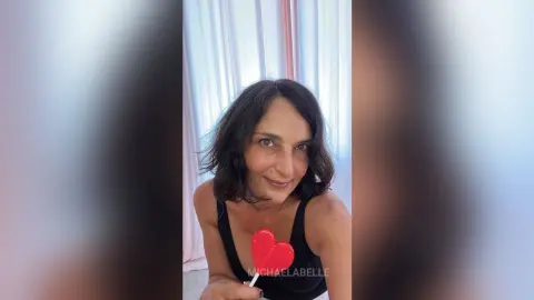 MichaelaBelle smiles while wearing a black tank top, holding a small red heart prop near her chest.