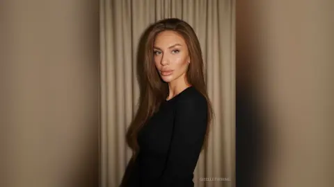 Elegant woman in a black long-sleeve top posing against neutral curtains in a moody, sophisticated setting.