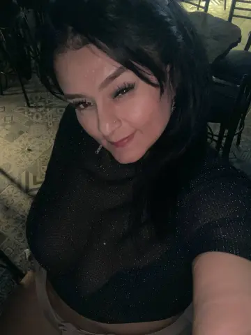 Smiling woman in a black glittery mesh top posing in a dimly lit restaurant setting.