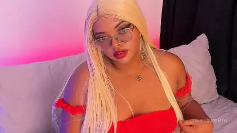 Blonde woman in a red top and glasses sitting on a bed with pink ambient lighting.