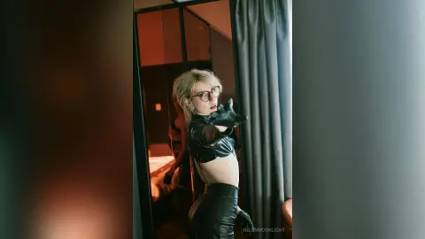 Stylish blonde woman in black leather outfit and glasses posing in a moody, modern interior setting.