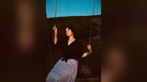 Young woman in a black top and long skirt sitting on a swing against a blue sky background.