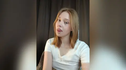 EvelinaMoorina is a blonde woman in a white top looking at the camera and licking her lips in front of dark curtains.