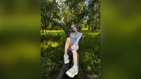 Young woman in a blue crop top and shorts sitting on a tree branch in a sunny green forest.
