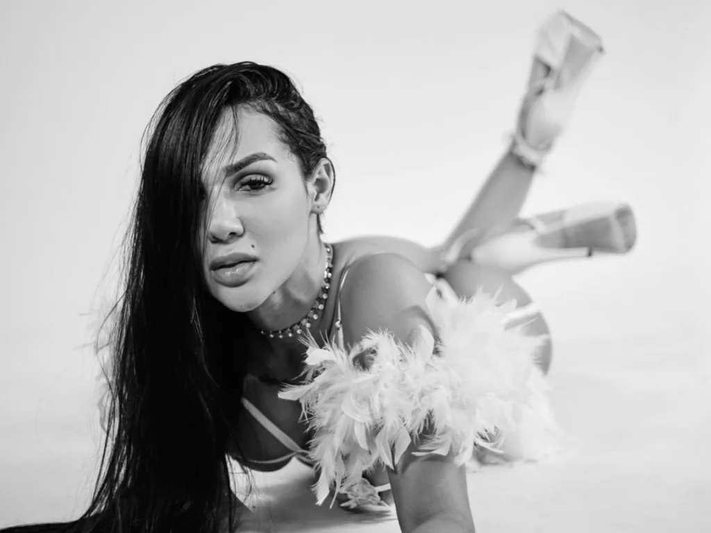 heracirce poses with long dark hair, wearing a black sheer mesh top with yellow caution tape patterns visible across her chest and shoulders.
