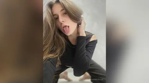 IngridBeckton poses with her long brown hair and tongue out, wearing a black long-sleeved crop top and black leggings.