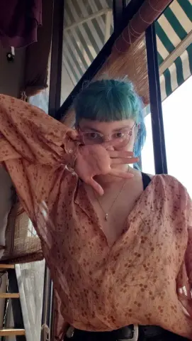 Woman with blue hair and glasses wearing a floral vintage blouse in a sunlit outdoor cafe setting.