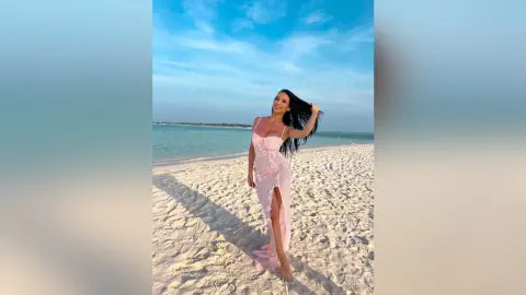 Smiling woman in a pink lace maxi dress standing on a sunny tropical beach under a blue sky.