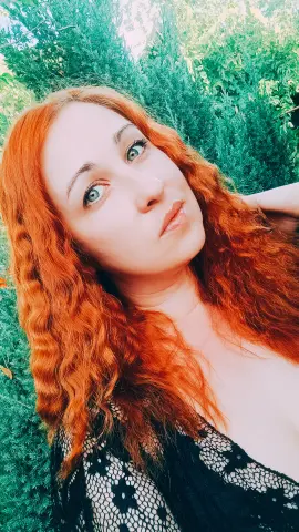 Jill_Stevens shows off her long, wavy red hair and light eyes while wearing a black lace top against a background of green leaves.