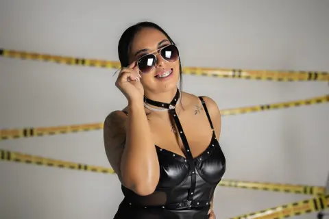 Smiling woman in black leather bodysuit and sunglasses posing against a white background with caution tape.